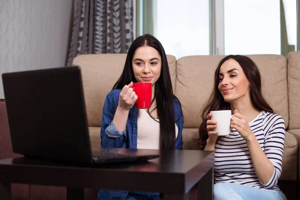ノート パソコンを自宅から映画を見て つの若い姉妹が床とコーヒーまたは紅茶の上に座ってください — ストック写真