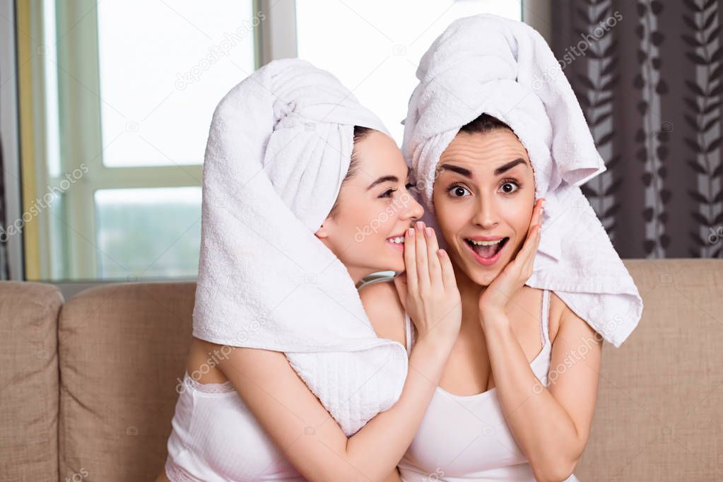 Beautiful smiling young sisters or girl friends in white towels on their heads after a spa gossip or discuss something sitting on the couch. The girls are resting.