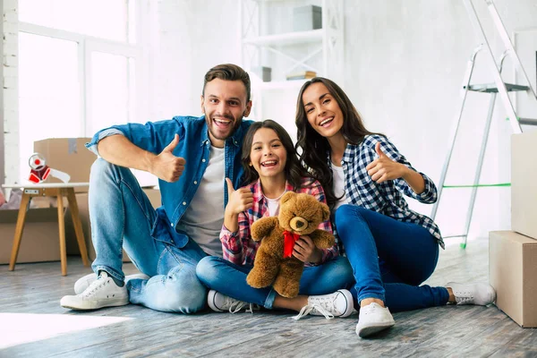 Looking forward to our bright future. Young father in denim outfit little teenage daughter and beautiful mother are sitting on the floor, hugging and smiling happily after moving house.