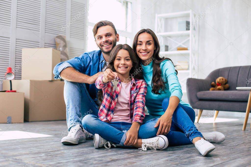 Looking forward to our bright future. Young father in denim outfit little teenage daughter and beautiful mother are sitting on the floor, hugging and smiling happily after moving house.