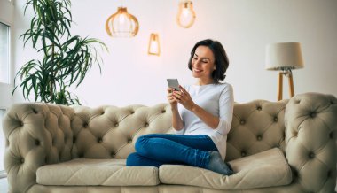Sıcak bir atmosfer. Gündelik giysiler içinde güzel bir kadın elinde bir akıllı telefon tutuyor ve yumuşak bir kanepede otururken gülümsüyor..