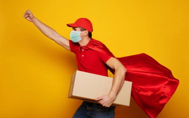 Tıbbi güvenlik maskeli genç teslimatçı sarı arka planda izole edilmiş bir kart kutusu taşıyor. Karantina döneminde internetten yiyecek alın..