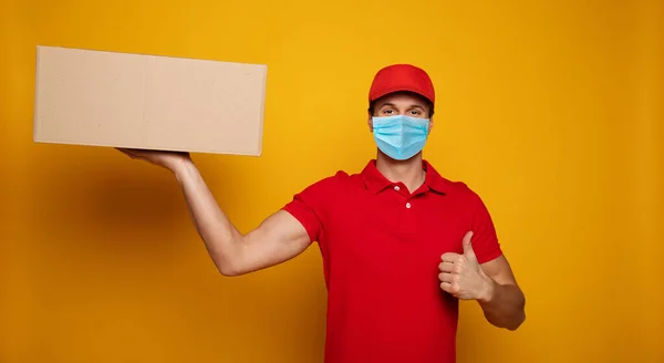 Young delivery man in medical safety mask holding and carrying a card box isolated on yellow background. Buy food online in the quarantine period.
