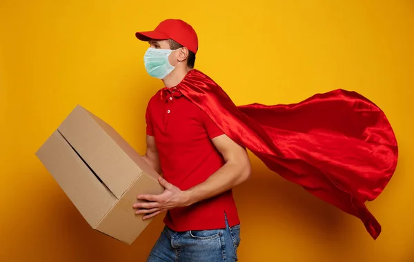 Young delivery man in medical safety mask holding and carrying a card box isolated on yellow background. Buy food online in the quarantine period.