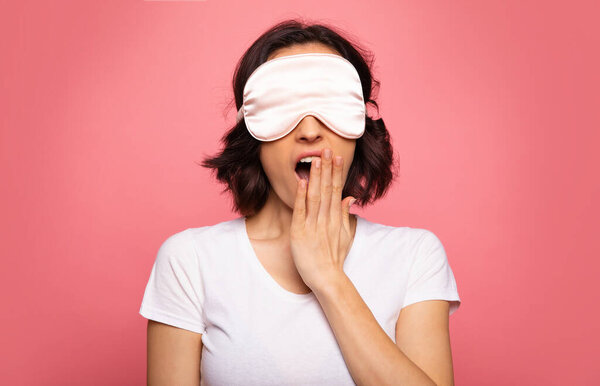 Craving for sleep. Close-up photo of a yawning young woman, who is wearing pajamas and a face mask.