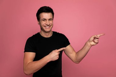 Terfi ettim. Siyah tişörtlü çekici bir erkeğin yakın plan fotoğrafı. Kameraya bakıyor, gülümsüyor ve iki eliyle de sağ elini gösteriyor..