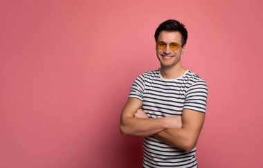 Çizgili ruh hali. Giysileri ve sarı gözlükleri olan genç ve güçlü bir adamın yakın plan fotoğrafı. Kollarını kavuşturup kameraya bakıyor..