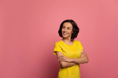 Pembe ve sarı. Sarı tişörtlü çekici bir kızın yakın plan fotoğrafı. Kollarını kavuşturup geniş bir gülümseme sergiliyor..