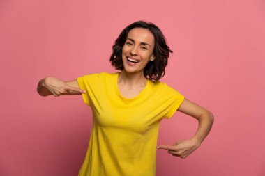 Logo için yer var. Sarı tişörtlü esmer kadının yakın plan fotoğrafı. Kameraya bakıyor, gülüyor ve göğsünü işaret ediyor..