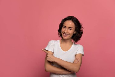 Basit bir güzellik. Mükemmel ciltli, beyaz tişörtlü güzel bir kadın kameraya bakıp gülümsüyor. Kolları kavuşturulmuş ve solunu işaret ediyor..