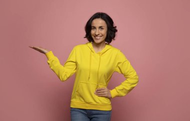 Genç yetişkin. Sarı kapüşonlu şık bir genç kızın yakın plan fotoğrafı, kameraya bakıyor ve gülümsüyor, sağ avucunu yukarı kaldırıyor ve sol elini kalçasında tutuyor..