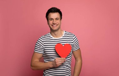 Ben de seni. Çizgili tişörtlü neşeli bir adamın yakın plan fotoğrafı. Kameraya bakıyor, gülümsüyor ve elinde kalp şeklinde bir kartpostal tutuyor..