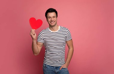 Genç aşık. Çizgili tişörtlü yakışıklı bir adamın yakın plan fotoğrafı. Kameraya bakıyor, gülümsüyor ve sağ elinde kağıt bir kalp tutuyor..
