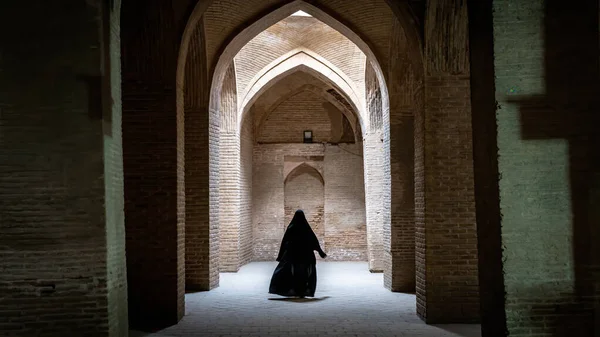Неопознанная иранская женщина в чёрном хиджабе идёт вдоль древних колонн мечети Джаме в Исфахане, Иран — стоковое фото