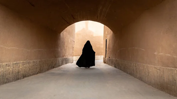 Azonosítatlan muszlim nő, fekete hidzsábba öltözve sétál a régi város, Yazd keskeny utcáján, Iránban.. — Stock Fotó