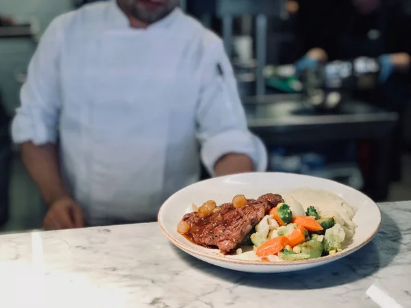 Chef Ready to Serve New York Steak with Shallot Onions and Vegetables at Restaurant Kitchen.