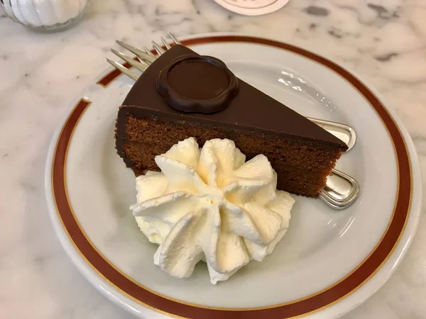 サッチャーケーキ 典型的なオーストリアのチョコレートケーキ チョコレートスポンジケーキの2つの厚板で構成されてい アプリコットジャムとホイップクリームの薄い層で区切られたプレート上 フォークで 食べる準備ができて — ストック写真