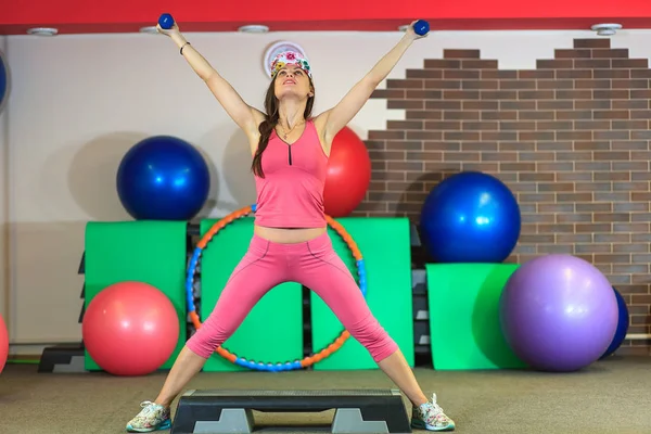 Menina branca bonita nova em um terno de esportes rosa faz exercícios físicos com halteres no centro de fitness . — Fotografia de Stock