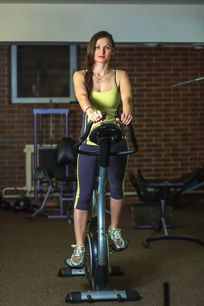 Young beautiful white girl in a yellow and gray sports suit is engaged on a stationary bike in the fitness club. — Stock Photo, Image