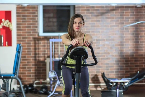 Young beautiful white girl in a yellow and gray sports suit is engaged on a stationary bike in the fitness club. — Stock Photo, Image