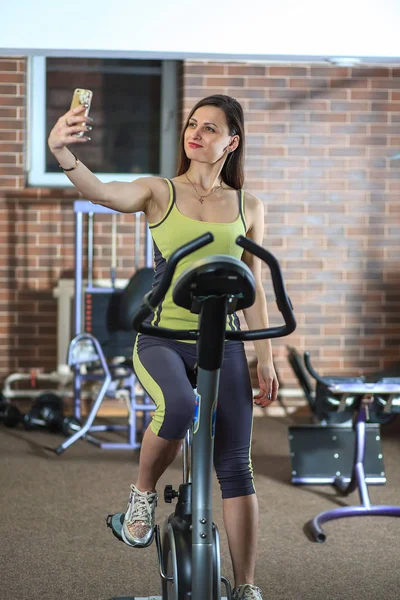 Genç güzel kız bir sarı beyaz ve gri spor takım elbise selfie fitness Club sabit bir bisiklet üzerinde var. — Stok fotoğraf