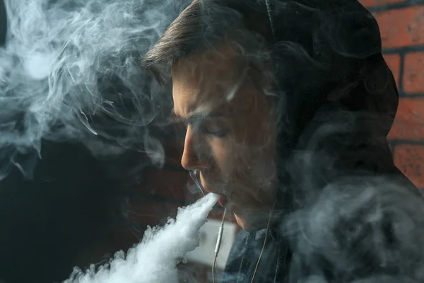 Young handsome white guy is admiting puffs of steam from the electronic cigarette. Vaping. Teenager listening to music. — Stock Photo, Image
