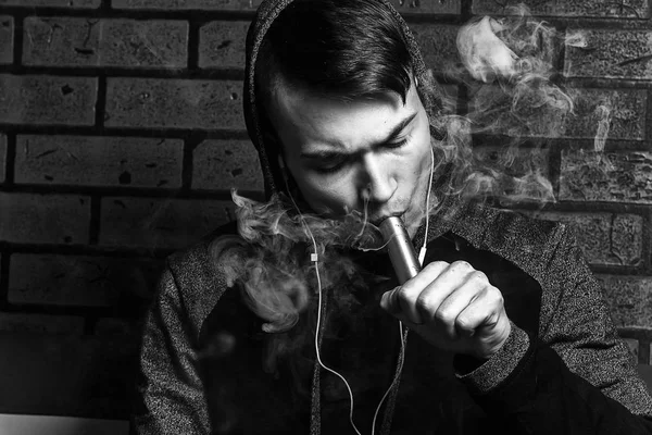 Jovem branco bonito está admitindo sopros de vapor do cigarro eletrônico. Vapor. Adolescente a ouvir música. Foto em preto e branco . — Fotografia de Stock