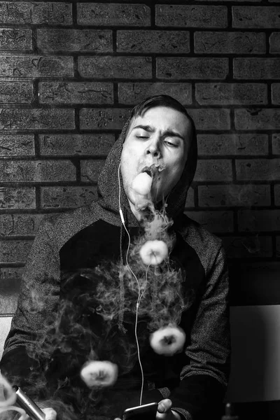 Der junge, gut aussehende weiße Mann bewundert die Dampfschwaden der elektronischen Zigarette. Dämpfen. Teenager hören Musik. Schwarz-Weiß-Foto. — Stockfoto
