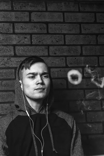 Young handsome white guy is admiting puffs of steam from the electronic cigarette. Vaping. Teenager listening to music. Black and white photo. — Stock Photo, Image