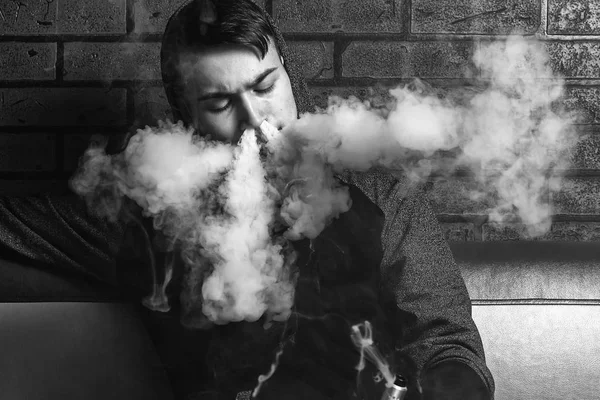Young handsome white guy is admiting puffs of steam from the electronic cigarette. Vaping. Teenager listening to music. Black and white photo. — Stock Photo, Image