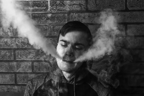 Jovem branco bonito está admitindo sopros de vapor do cigarro eletrônico. Vapor. Adolescente a ouvir música. Foto em preto e branco . — Fotografia de Stock