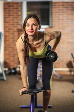 Genç güzel kız bir sarı beyaz ve gri spor takım egzersizler fitness kulübü eğitim cihaz üzerinde bir dumbbell ile yapıyor.