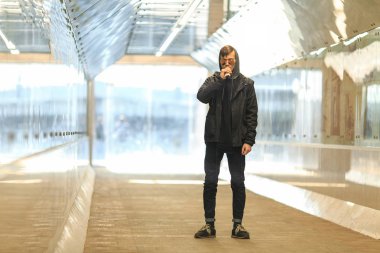 Vape. Yakışıklı genç bir beyaz adam güneş gözlüğü ve başlık ayakta ve yeraltı geçidi içinde elektronik sigara sigara.