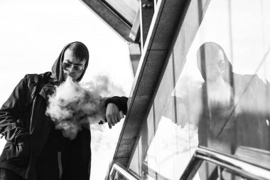 Vape. Yakışıklı genç bir beyaz adam güneş gözlüğü ve başlık ayakta ve güneşli bir günde köprünün iniş yakınındaki elektronik sigara sigara. Siyah ve beyaz fotoğraf.