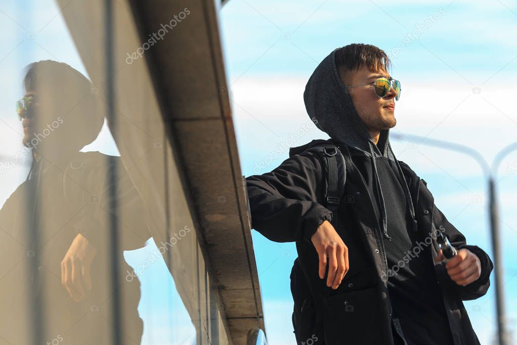 Vape. A handsome young white guy in sunglasses and in the hood is standing and smoking an electronic cigarette near the descent into the underpass on a sunny day.