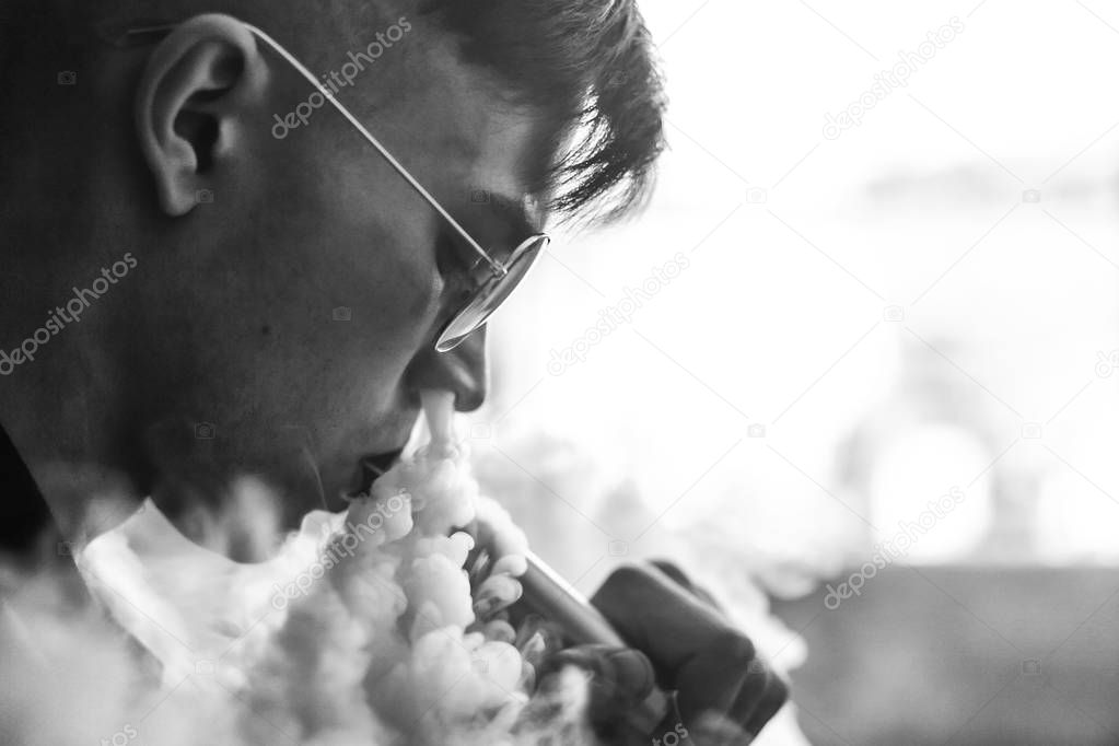 Vape. Young handsome white guy in sunglasses is admiting puffs of steam from the electronic cigarette. Vaping. Teenager listening to music. Black and white photo.