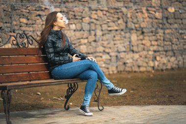 Vape. Genç, güzel ve beyaz bir kız vintage bir bankta oturan ve elektronik sigara sigara içiyor. Yaşam tarzı.