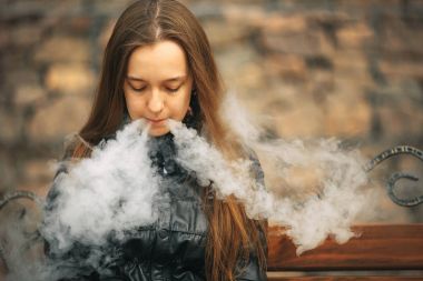 Vape. Genç, güzel ve beyaz bir kız vintage bir bankta oturan ve elektronik sigara sigara içiyor. Yaşam tarzı.