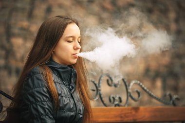 Vape. Genç, güzel ve beyaz bir kız vintage bir bankta oturan ve elektronik sigara sigara içiyor. Yaşam tarzı.