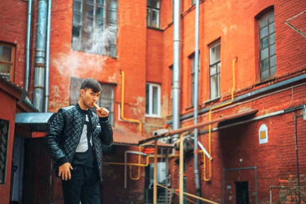 Vape. A handsome young white guy blows steam from an electronic cigarette in a vintage old red yard. Vaping. — Stock Photo, Image