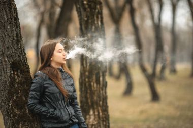 Vape. Genç, güzel ve beyaz bir kız ağaca yakın elektronik sigara içiyor. Yaşam tarzı.