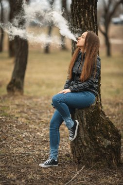 Vape. Genç, güzel ve beyaz bir kız ağaca yakın elektronik sigara içiyor. Yaşam tarzı.