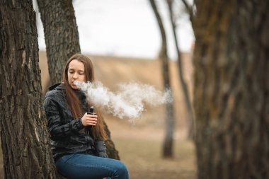 Vape. Genç, güzel ve beyaz bir kız ağaca yakın elektronik sigara içiyor. Yaşam tarzı.