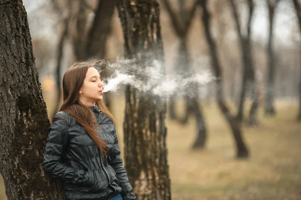 Vape. Młoda piękna dziewczyna białe dymy elektroniczny papieros w pobliżu drzewa. Styl życia. — Zdjęcie stockowe