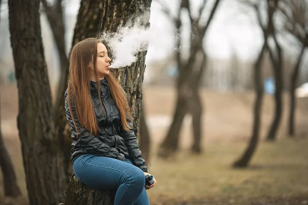 Вэйп. Молодая красивая белая девушка курит электронную сигарету возле дерева. Стиль жизни . — стоковое фото