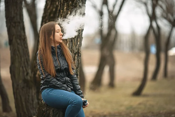 Vape. Genç, güzel ve beyaz bir kız ağaca yakın elektronik sigara içiyor. Yaşam tarzı.