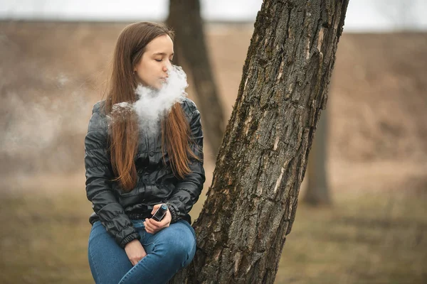 Vape. Młoda piękna dziewczyna białe dymy elektroniczny papieros w pobliżu drzewa. Styl życia. — Zdjęcie stockowe