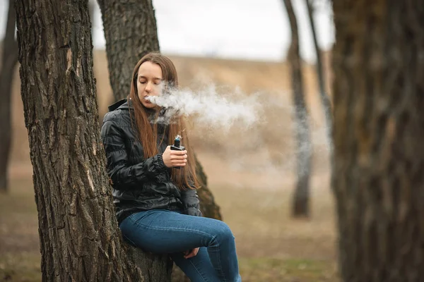 Vape. Een mooie witte meisje rookt een elektronische sigaret in de buurt van een boom. Levensstijl. — Stockfoto