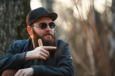 Vape. Acımasız bir genç adam güneş gözlüğü ve bir kap içinde büyük sakallı ve gün batımında ormanda elektronik sigara ile. Yaşam tarzı.