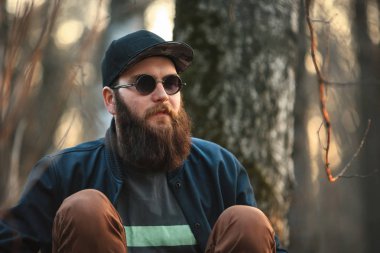 Güneş gözlüklü büyük sakal ve gün batımında ormanda bir kap ile acımasız bir genç adam. Yaşam tarzı.